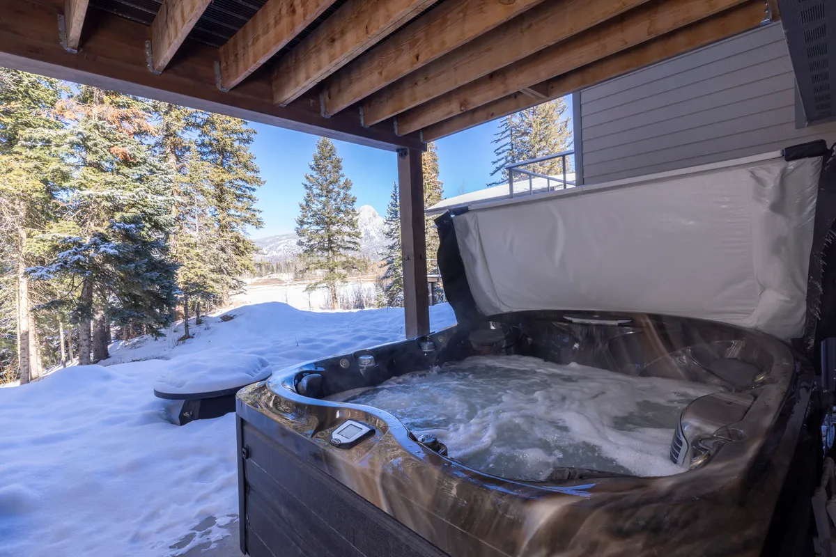 A steaming hot tub sits in the snow, surrounded by pine trees and overlooking a breathtaking mountain vista. The scene captures the serene luxury of unwinding after a day’s adventure, blending relaxation with spectacular winter scenery.