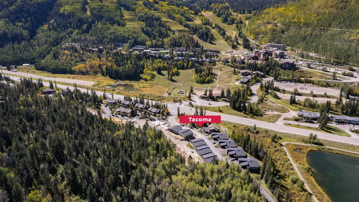 The image showcases the inviting exterior of Tacoma Vista 115, nestled among lush trees with a clear view of majestic mountains in the backdrop. The Tacoma sign welcomes guests at the entrance, while the nearby lake and scenic road hint at outdoor adventures just steps away. The setting exudes tranquility and a strong connection to nature, with vibrant, seasonally changing foliage enhancing the picturesque resort environment.