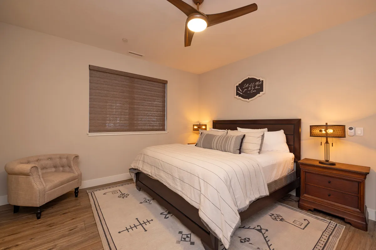 A serene bedroom scene with a bed adorned in striped bedding, a nightstand with a soft-glowing lamp, and a comfortable chair nestled in the corner. Artistic wall decor and gentle lighting create a peaceful, restful vibe ideal for unwinding.