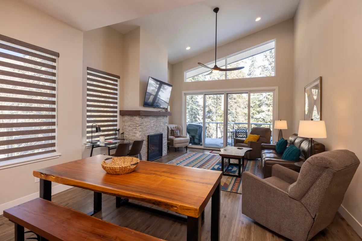 This image showcases an inviting open-concept space featuring a dining table set for gatherings, a cozy fireplace, large windows overlooking natural scenery, and plush, comfortable seating. The layout connects the living and dining areas, highlighting the welcoming atmosphere and the focus on togetherness.