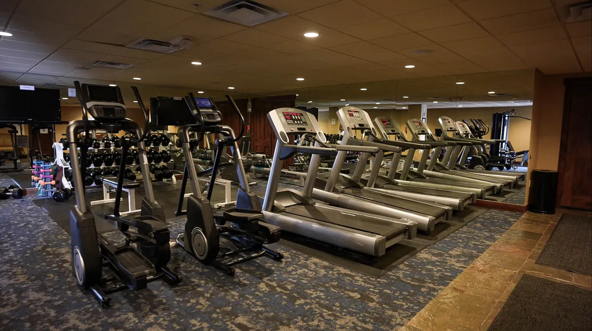 This modern fitness room features elliptical machines, treadmills, dumbbells, and exercise mats arranged against mirrored walls. The space is brightly lit and designed for both solo workouts and group fitness activities.