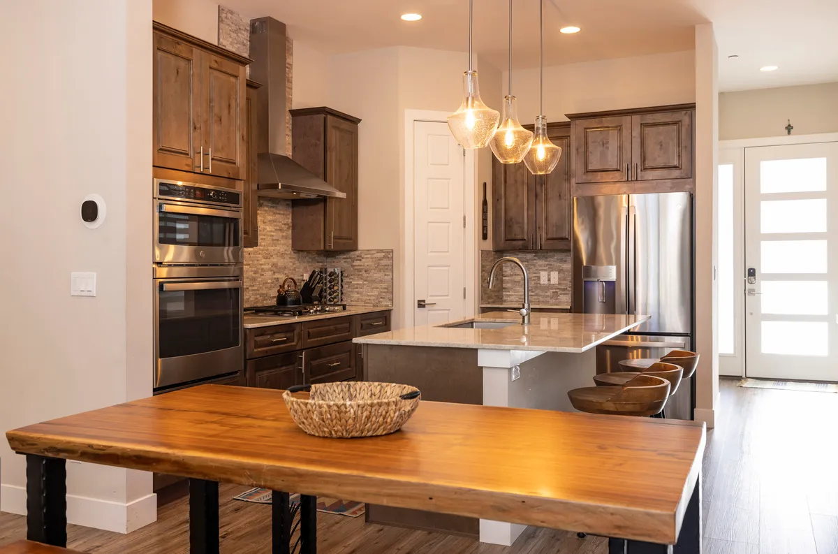 A bright, modern kitchen and dining area featuring a spacious kitchen island, stainless steel appliances, and a dining table set under hanging light fixtures. The open-plan design invites guests to gather, cook, and dine together in a welcoming space.