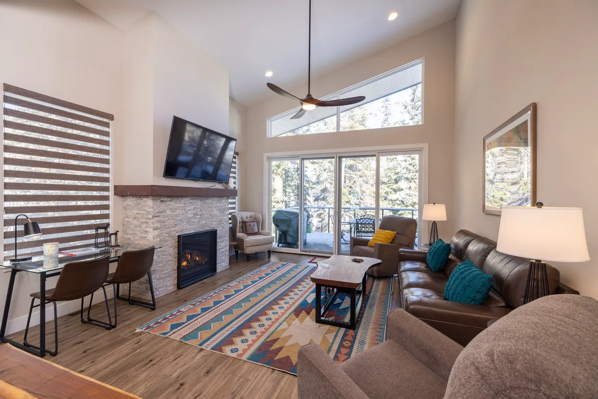 The photo offers a broad view of the living and dining area, with a prominent fireplace, expansive windows framing mountain vistas, a cozy sofa, a soft area rug, and a dining table. The natural light highlights the warmth and comfort of the communal space.