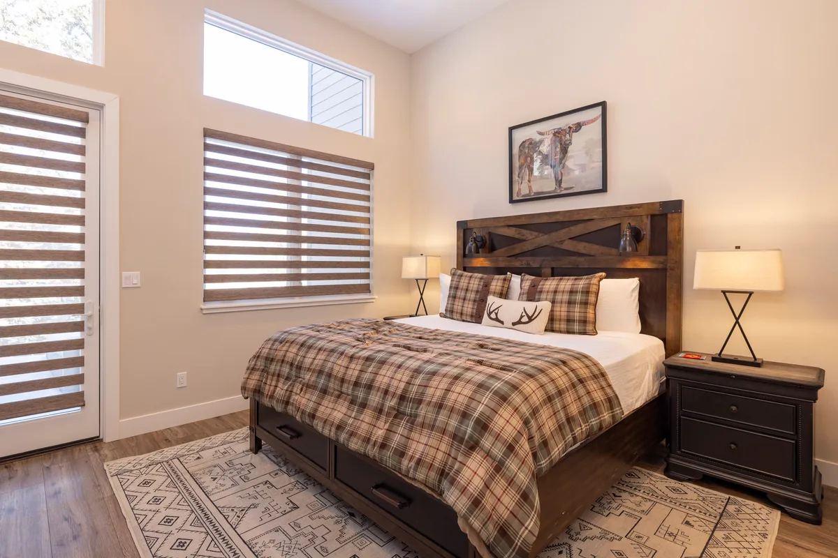 A warm, inviting bedroom featuring a wooden bed frame with plaid bedding, complemented by tasteful artwork on the wall and cozy nightstands with lamps. Soft natural light filters in, creating a peaceful atmosphere for relaxation or a good night's sleep.
