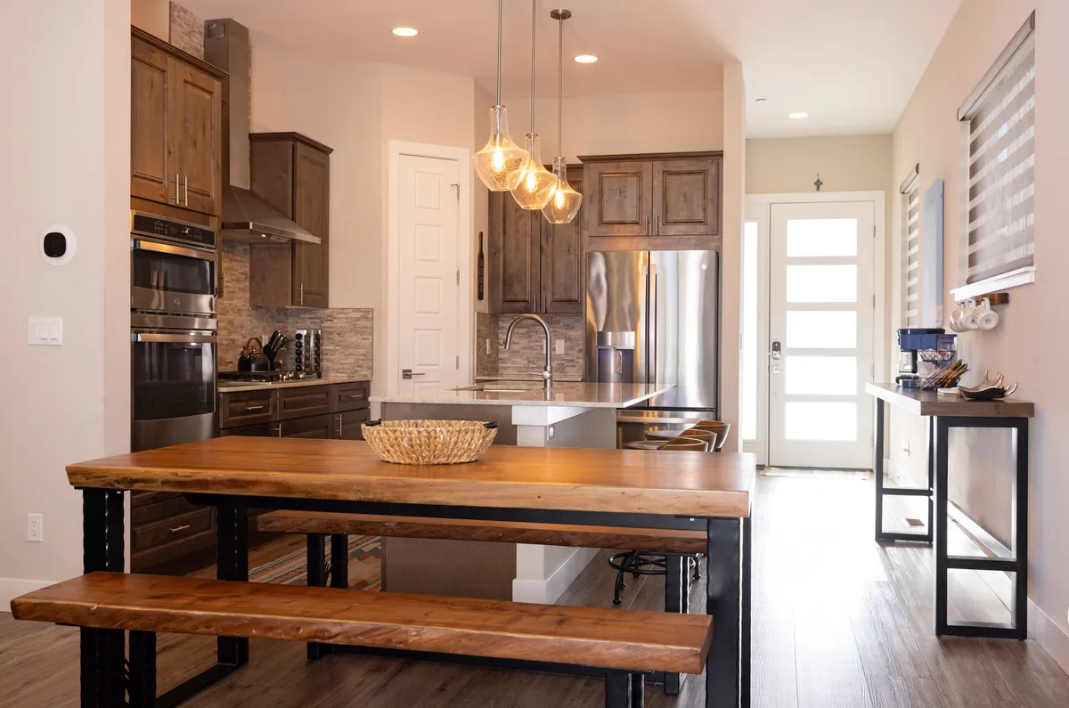 A wide view of a chef’s kitchen and communal dining area, featuring a wooden dining table under stylish pendant lights, a central kitchen island, and gleaming stainless steel appliances. The open layout allows the kitchen to flow seamlessly into the dining space, evoking a sense of warmth and shared activity. Family members can be visualized gathering for meals or casual coffee conversations.