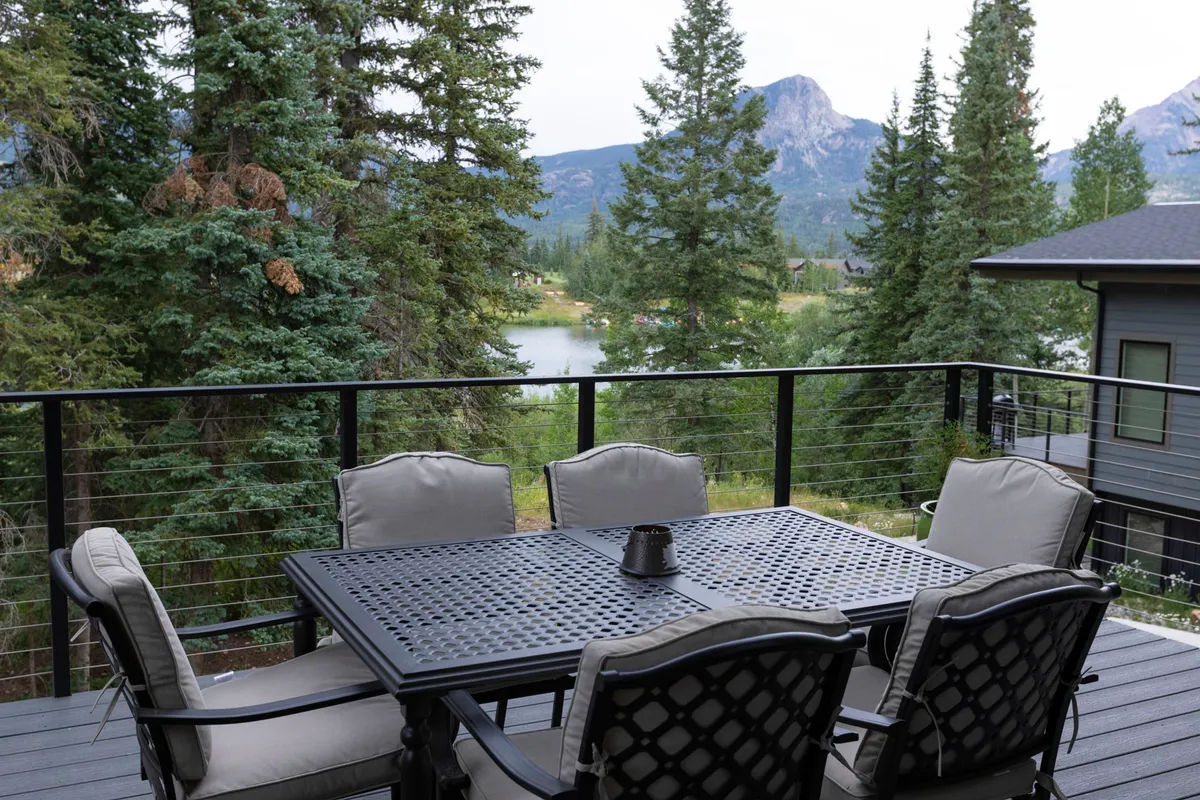 An inviting exterior shot of a dining table set on a patio, overlooking a tranquil lake surrounded by trees and backed by scenic mountains. The setting sun casts a warm glow, making it an ideal spot for outdoor dining and sunset barbecues.