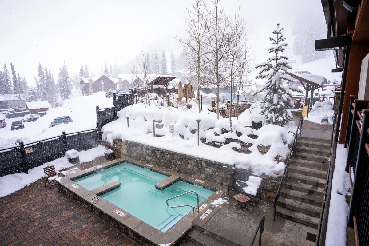 A serene outdoor hot tub set amidst a snow-covered landscape, with surrounding trees and steps leading up to the spa area. The scene radiates tranquility, inviting guests to relax while soaking in the warmth and admiring the beautiful winter views.