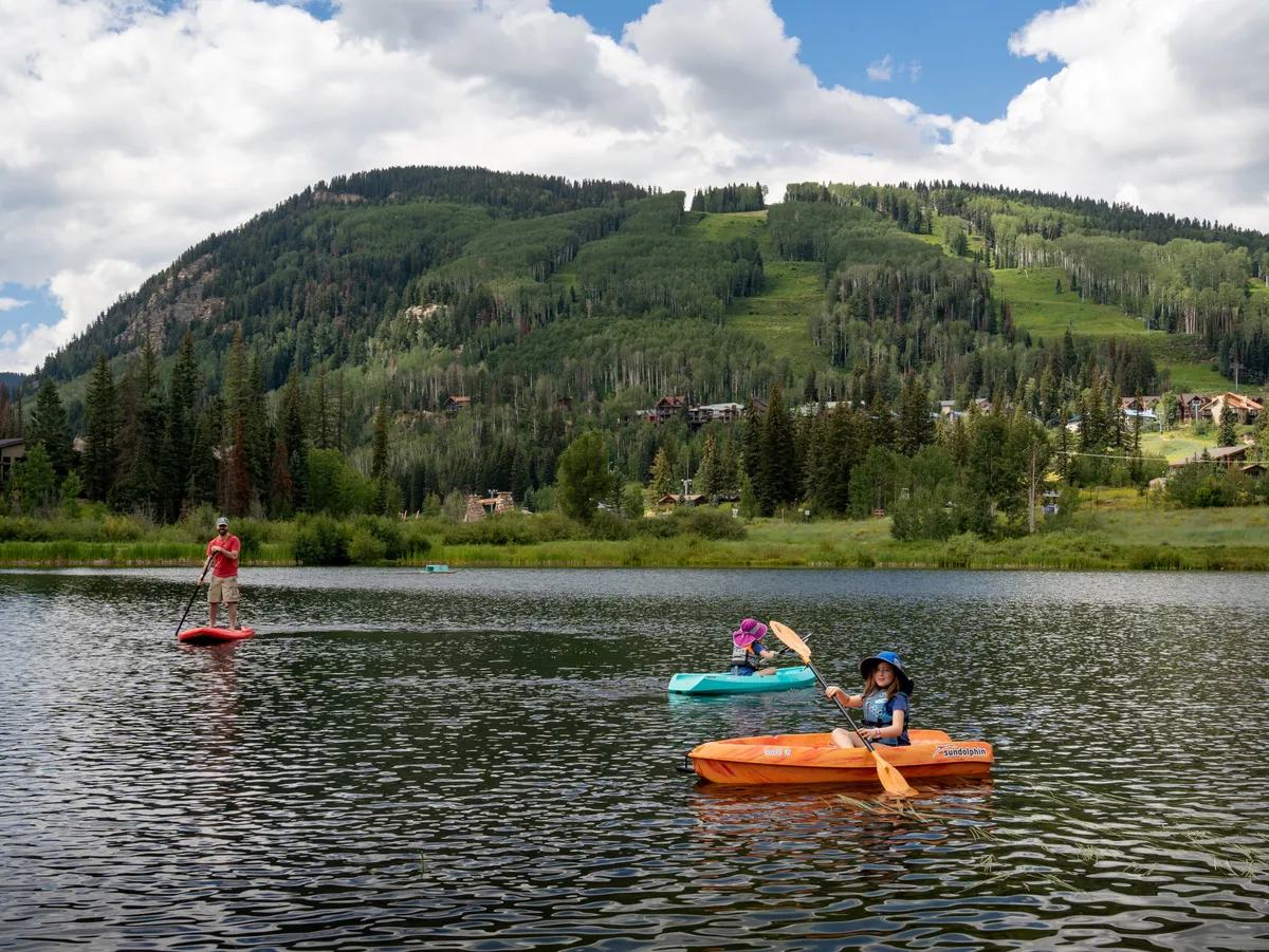 A lively lakeside scene showcasing a paddleboarder gliding across calm waters, with kayakers nearby and majestic mountains reflected on the lake's surface. The sunlight enhances the playful and relaxed mood, emphasizing outdoor recreation and family bonding.